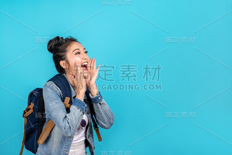 快乐的亚洲女人旅行背包客大声呼喊着在蓝色背景上的copyspace。可爱的亚洲女孩微笑着穿着休闲牛仔