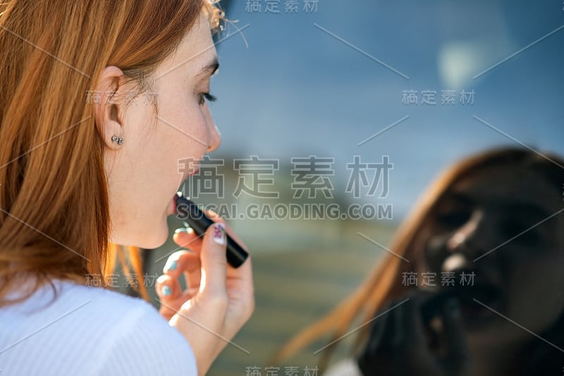 一个年轻的红发女司机的特写纠正她的化妆用深红色口红看在汽车反光窗口。