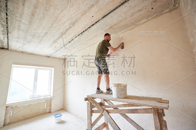 一个年轻人正站在木架上，用抹灰铲在靠窗的墙上工作。