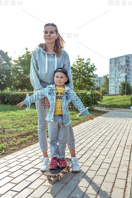 幸福年轻的家庭，女人妈妈带着孩子男孩，滑板，育儿帮助支持，育儿，第一次滑板课。在夏天的城市。笑的情绪