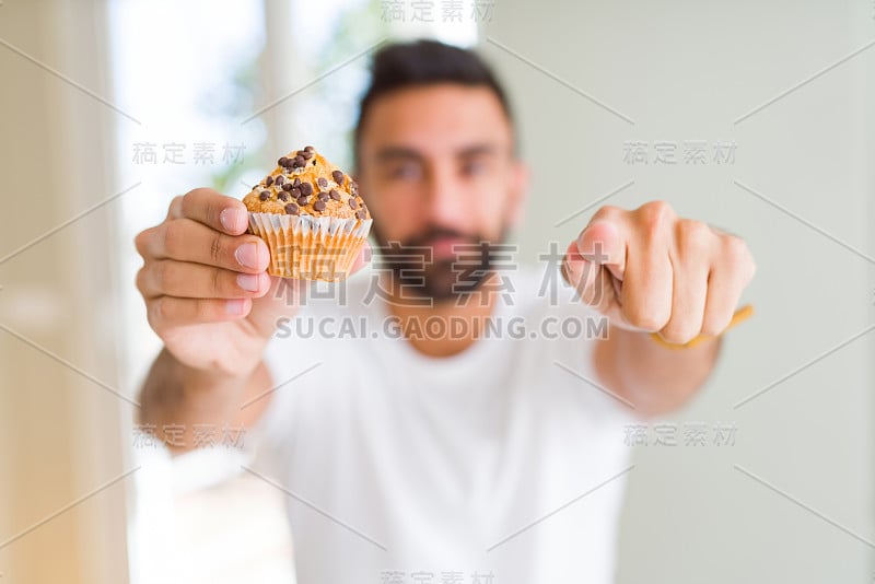 英俊的西班牙裔男子吃着巧克力片松饼，手指对着镜头和你，手势，正面自信的手势