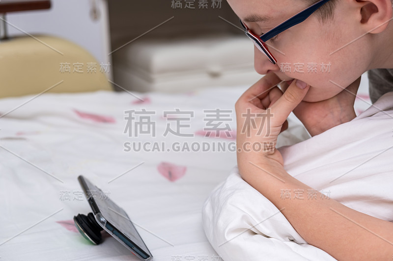 一个戴眼镜的十多岁的男孩躺在床上用智能手机看视频