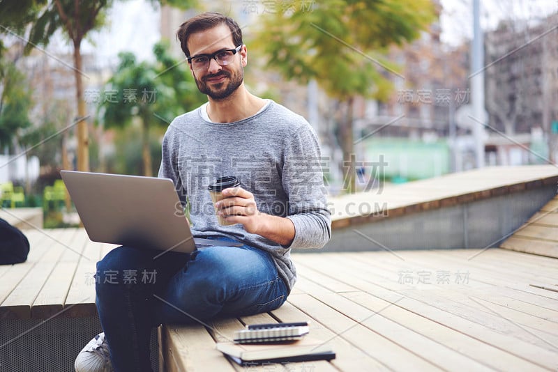 这是一幅漂亮的留着胡子的学生戴着眼镜坐在户外，手提电脑连接到高速4G网络，手里拿着美味的咖啡，微笑着