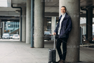 时尚成熟的游客站在户外。一个提箱子的男人斜靠在墙上，看向别处。