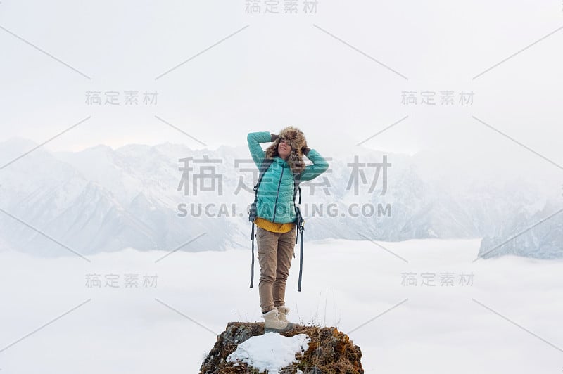 一个快乐微笑的嬉皮士女孩旅行，穿着羽绒服，背着背包，戴着一顶大毛皮帽子站在岩石上