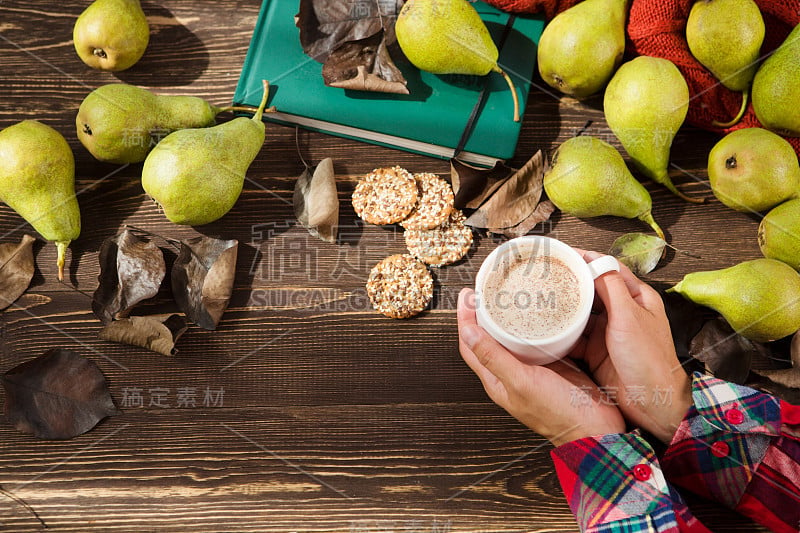 女性手捧热可可或巧克力杯，用梨、饼干、树叶放在旧木桌上。秋天的成分。