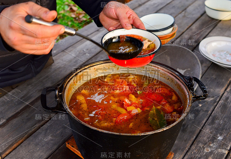 在大自然中烹饪食物。男人们，厨师从锅里倒一勺汤到鹤碗里