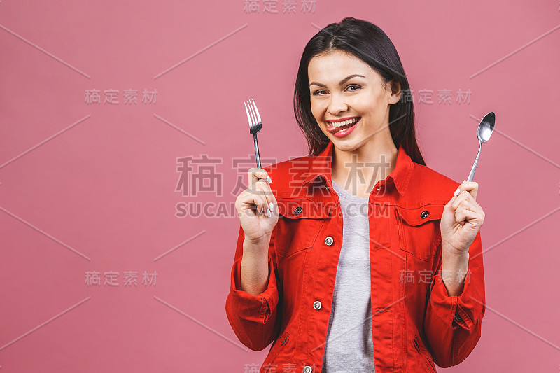 吃晚饭了!年轻美丽的女人的肖像穿着红色休闲衬衫，拿着勺子和叉子孤立在粉红色的背景。