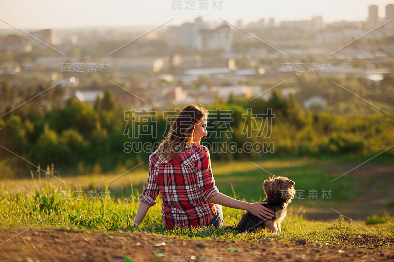 穿着红色格子衬衫的年轻女子和她的宠物约克夏犬坐在公园的草地上。朋友们看着城市