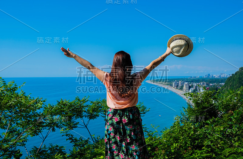 女孩肖像草帽，珊瑚色衬衫和热带印花裙。眺望大海和全景城市。后视图。