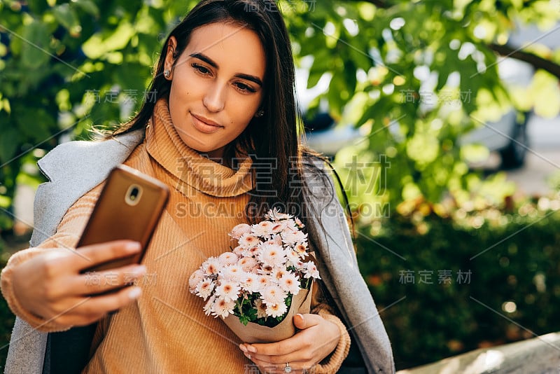 漂亮的女孩说话的自画像与一束花在手机户外反对在公园的绿色植物。美丽的年轻女性站在城市的街道上用她的设