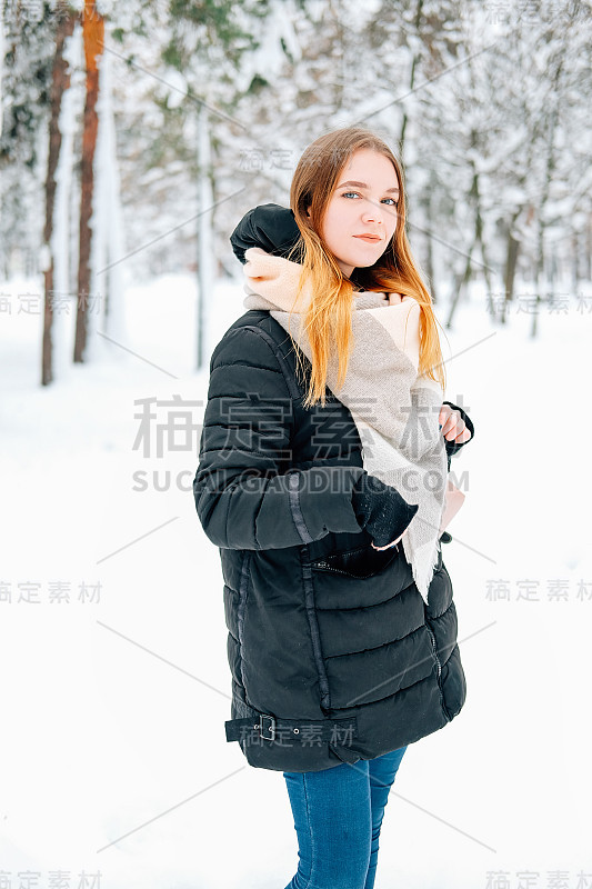 迷人的金发年轻女子走在冬天的森林里满是雪，穿着黑色皮大衣，牛仔裤，粉色皮靴，背包和米色披肩的休闲服装