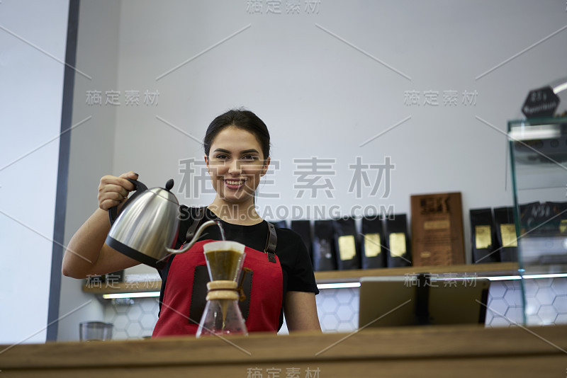 正面的女咖啡师微笑着看着镜头制作美味的咖啡。漂亮的女服务员手里拿着水壶，在酒吧里为准备浓咖啡倒热水