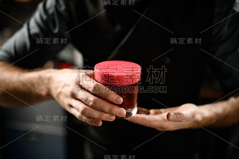 调酒师在用光滑的粉红色浮渣装饰的玻璃杯中端上金黄色的鸡尾酒