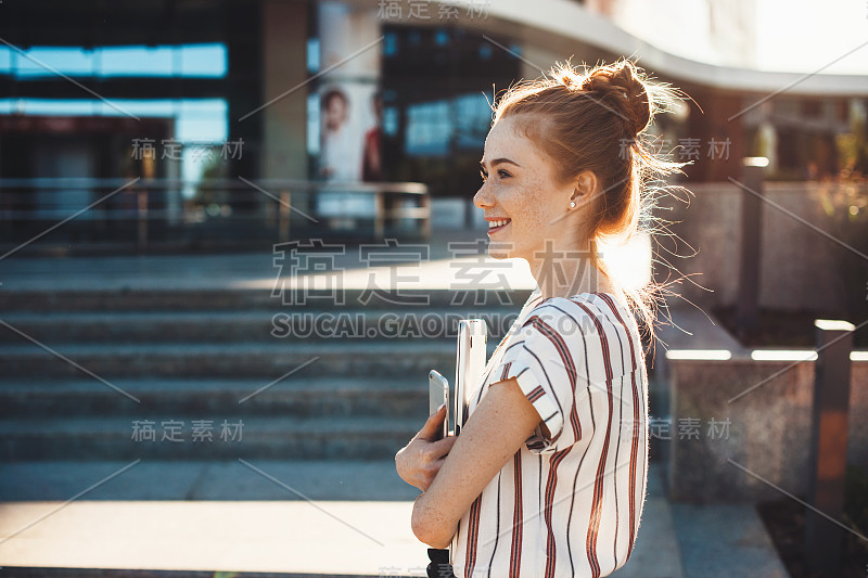 一个长着红头发和雀斑的开朗的白人女孩正在一座建筑前拿着一块平板看别处