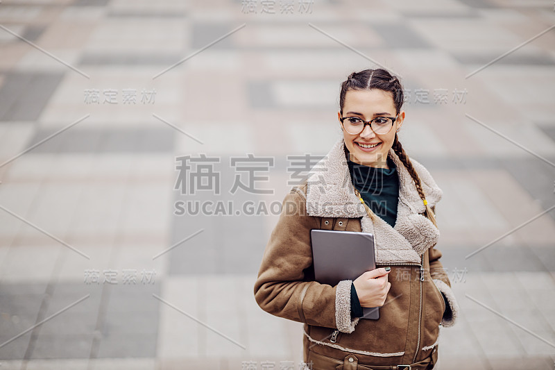 年轻女孩微笑着站在户外，拿着平板电脑，看着别处。全球通信和新技术概念。