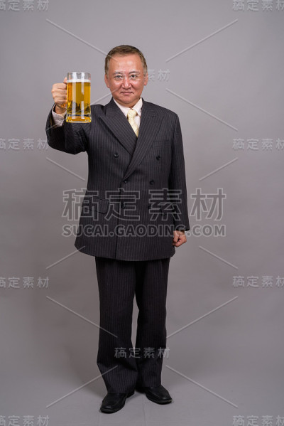 在灰色的背景下，一个成熟的亚洲商人拿着一杯啤酒