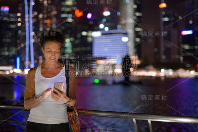 在新加坡的滨海湾，一个成熟的女人在晚上使用手机和短信
