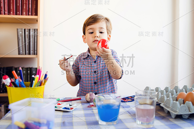 白种人小男孩两岁，拿着用水彩笔画的复活节彩蛋，站在家里的桌子旁为节日装饰