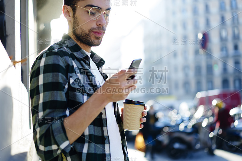 严肃英俊的男子戴着眼镜，站在阳光明媚的城市环境中，通过电话仔细观看有趣的4G网络视频。旅客休息与现代