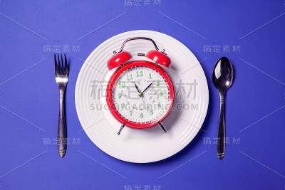 闹钟放在白色盘子上，勺子和叉子放在彩色背景上。饮食概念