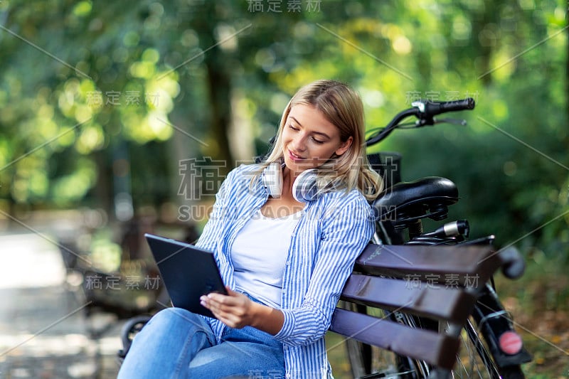 金发女人坐在长椅上，手上拿着平板电脑，脖子上戴着耳机