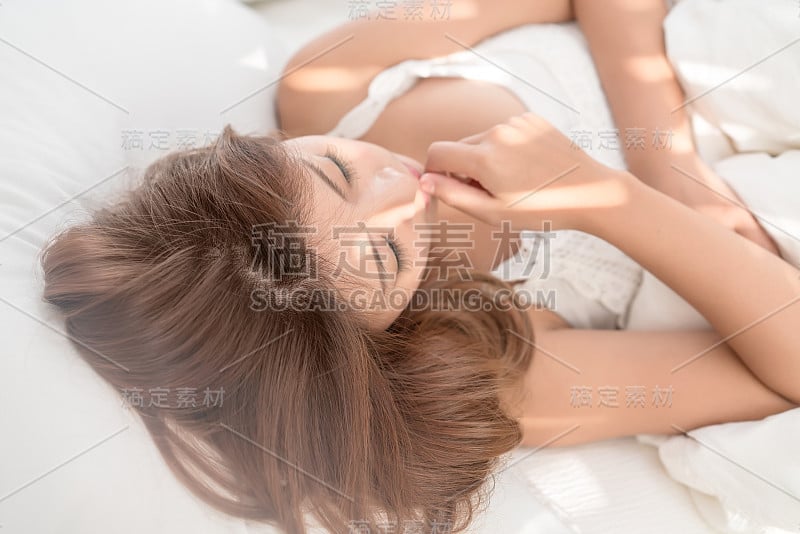 美丽的亚洲女人肖像与迷人的微笑享受新鲜柔软的床上用品亚麻布床垫在白色的卧室现代公寓。可爱的亚洲女孩睡