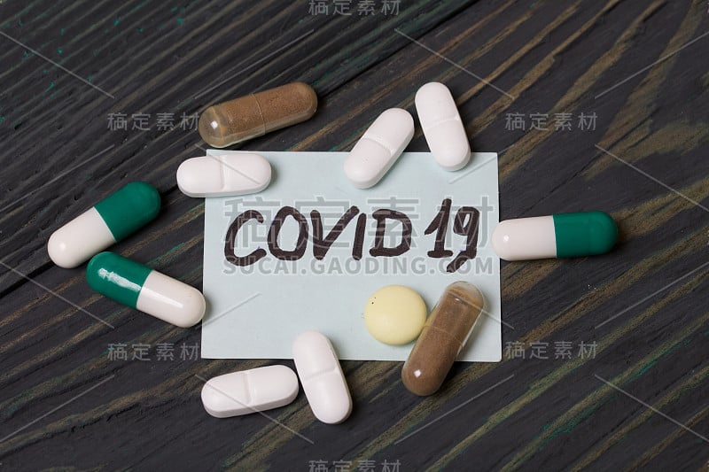 贴有COVID-19字样的贴纸。旁边是几颗形状和颜色各异的药丸。躺在刷过的松木板上。