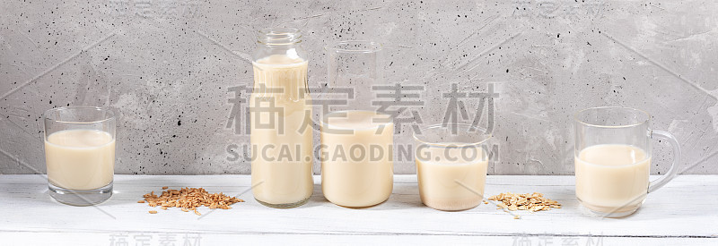 在灰色混凝土墙背景的白色木桌上，一排玻璃容器和燕麦牛奶的全景图像。