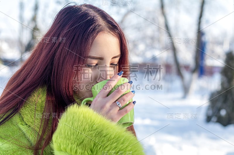 美丽可爱漂亮的红发女青少年在绿色毛皮大衣喝咖啡在阳光明媚的冬季城市公园。