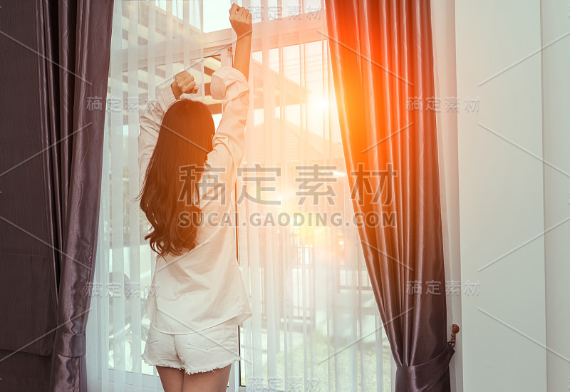 女人早上醒来，打开窗户后，在床上放松美好的一天在假日与阳光，在生活的家庭生活方式的后视图