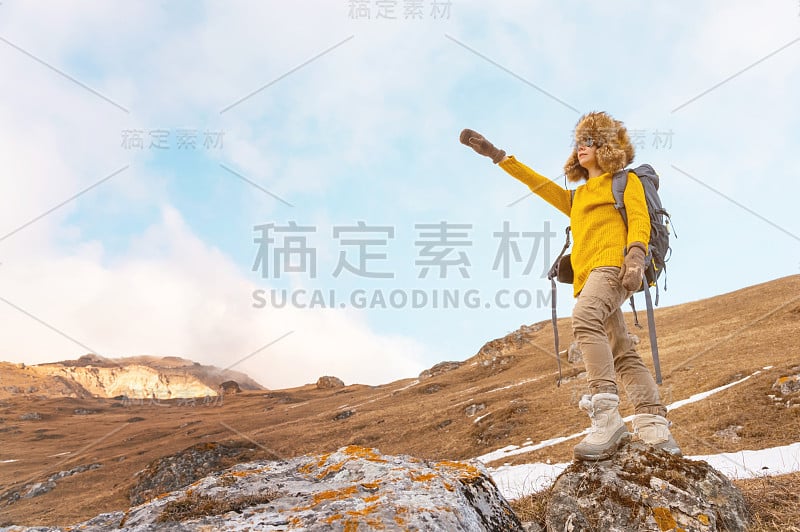 背包女孩戴着墨镜，戴着一顶大大的北方皮草帽，背上背着一个背包，指着一块岩石，看着隐藏在岩石中的悬崖