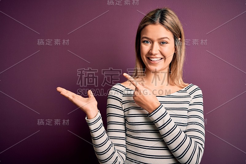 年轻美丽的金发女人穿着休闲条纹衬衫在紫色孤立的背景惊讶和微笑的相机，同时提出与手指。