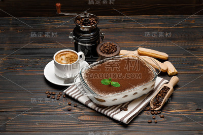传统的意大利提拉米苏甜点在玻璃烘焙盘，咖啡研磨机，savoiardi饼干和一杯新鲜的热咖啡在木背景