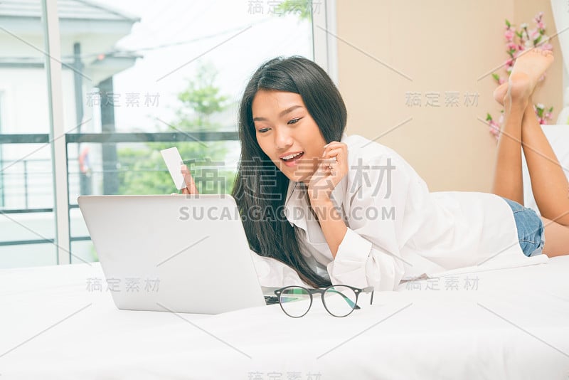 快乐随意美丽的亚洲女人坐在家里的床上用笔记本电脑工作。