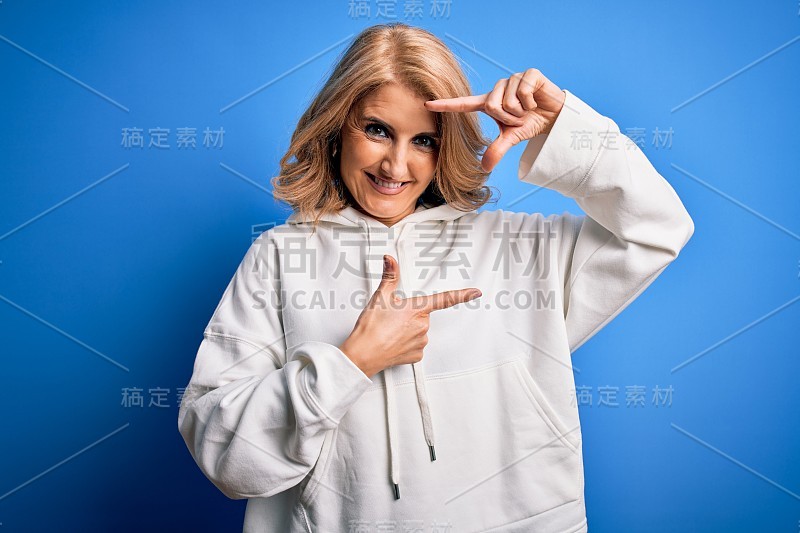 中年美丽的金发女运动员穿着运动服在孤立的蓝色背景微笑使框架与手和手指与幸福的脸。创意和摄影概念。