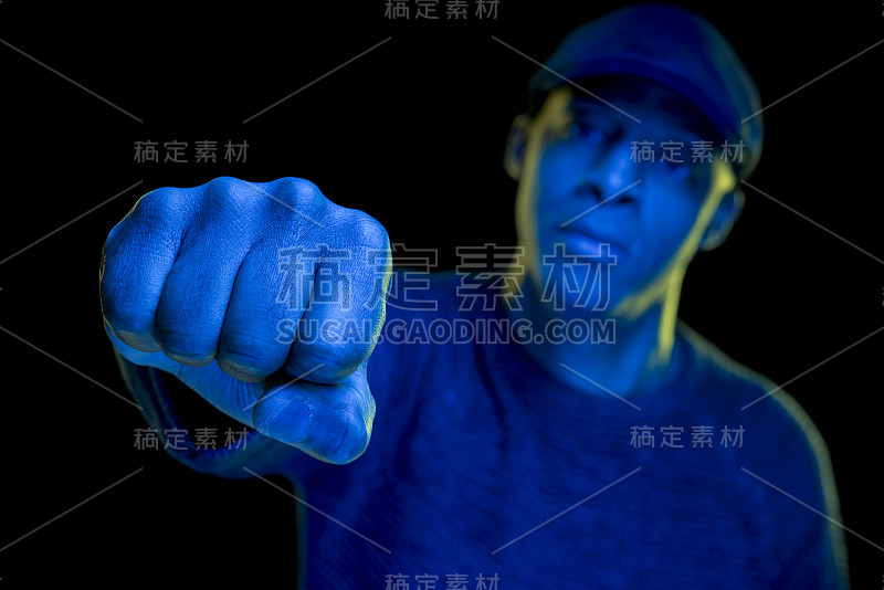 一个拳头的特写。黑衣人带着黄蓝相间的光，戴着一顶平帽，看着摄像机。