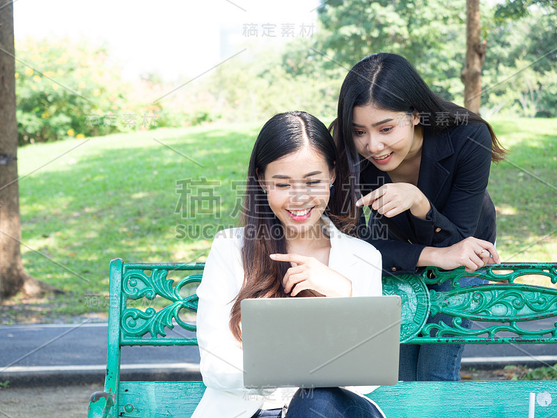 两个亚洲商业女人与电脑笔记本在公园工作。美女打字键盘笔记本空白屏幕和朋友站在后面与拷贝空间。