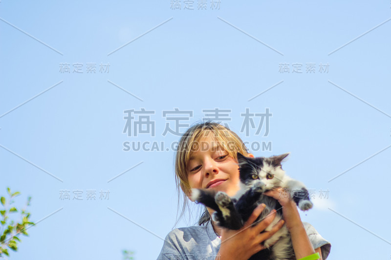 散焦金发微笑少女抱着白色和黑色的猫，小猫在蓝天的背景。户外,外面。宠物，爱护动物的概念。的焦点