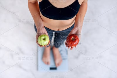 身材完美的运动女性拿着红绿苹果用电子秤测量体重。