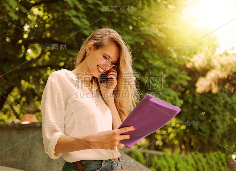 一个美丽的女商人的肖像，穿着休闲风格的衣服拿着一个文件夹的文件，并在户外走路打电话