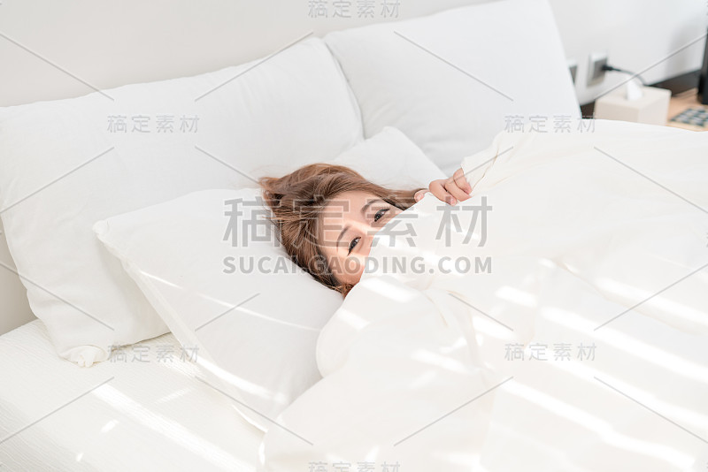 美丽的亚洲女人肖像与迷人的微笑享受新鲜柔软的床上用品亚麻布床垫在白色的卧室现代公寓。可爱的亚洲女孩睡