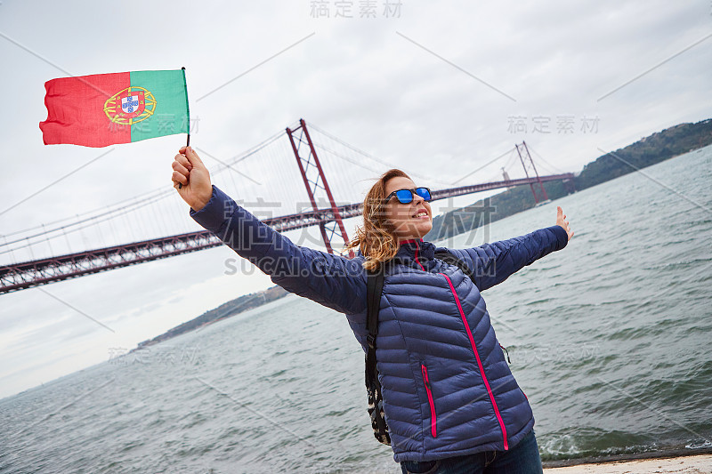 4月25日，年轻的女游客手持葡萄牙国旗，在著名的铁桥上欣赏风景，站在里斯本市的河边