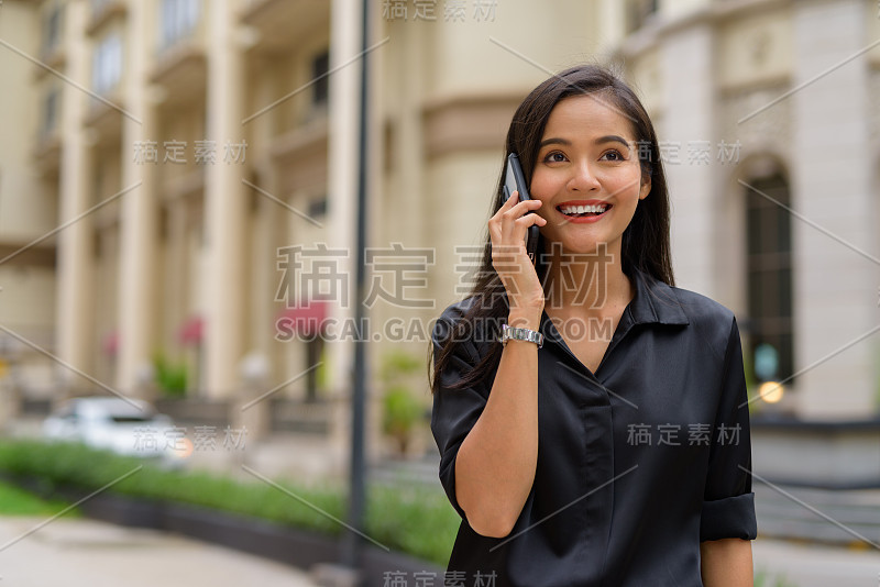 快乐的亚洲女商人在户外城市街道上打电话，而微笑和行走