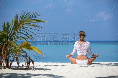 年轻健康的女人穿着白色衣服在海滩上练习瑜伽。马尔代夫