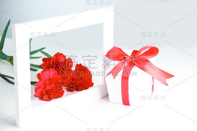 三朵红色康乃馨，白色包装的礼物，一个红色的蝴蝶结和框架在白色的背景。母亲节贺卡，国际妇女节。