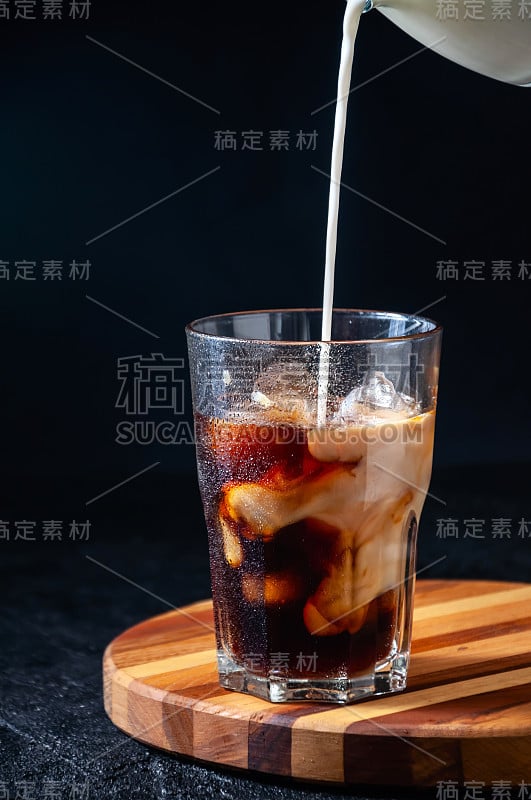 在黑色背景下，牛奶被倒入高杯冰咖啡中。概念清凉夏日饮品