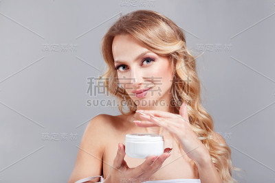 美丽的金发女子，有着清澈的皮肤和蓝色的眼睛，双手保持白色的罐子和奶油