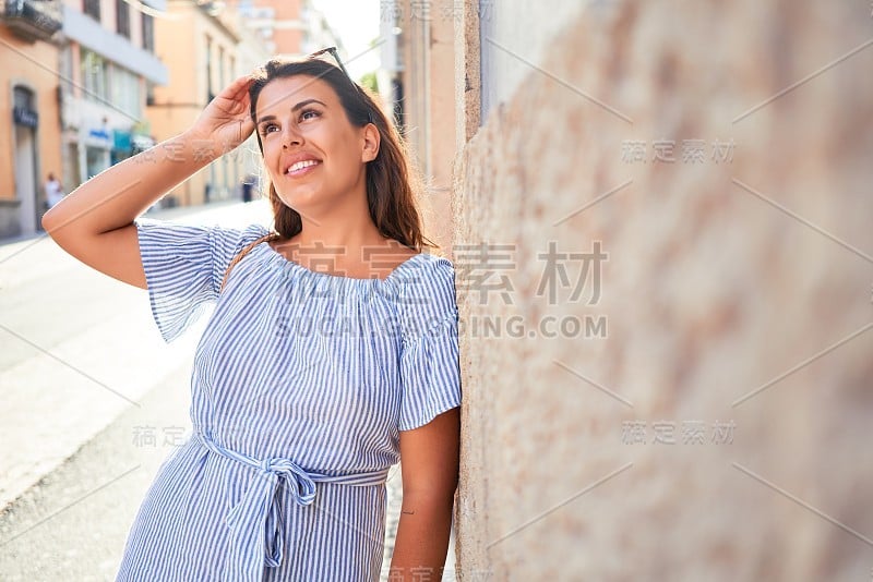 在一个阳光明媚的夏日，年轻美丽的女子靠在城市街道的墙上，快乐地微笑着