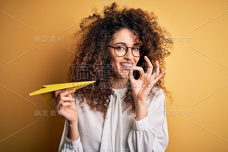 漂亮的卷发和穿洞的女人拿着纸飞机在黄色的背景做ok手势，优秀的象征